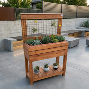 Un bac à fleurs surélevé en bois brun avec un treillis métallique, placé sur une terrasse moderne en béton. Le bac contient des plantes grasses et des plantes grimpantes qui s'enroulent autour de structures décoratives en acier.