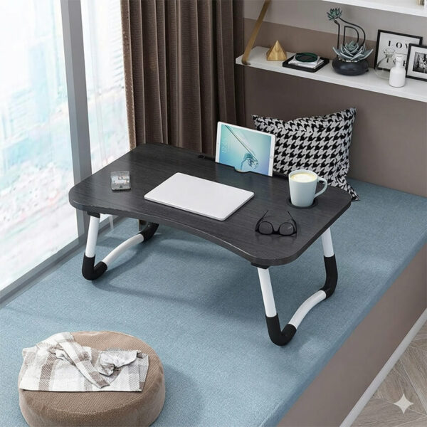 Table de lit pliante noire et blanche installée sur un canapé ou un lit confortable en Tunisie, portant un ordinateur portable ouvert, une tasse de café et une tablette avec des écouteurs posés à côté. Un moment de pure détente.